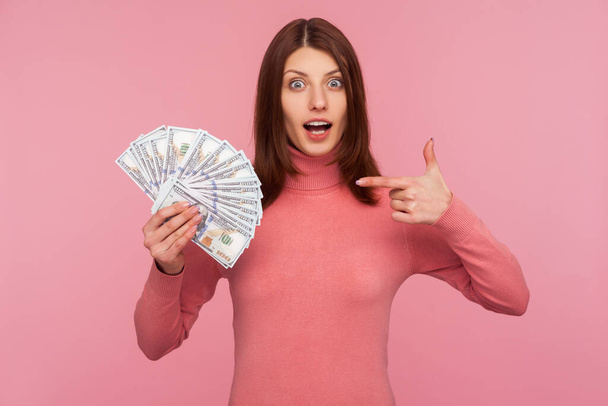 Surprised amazed woman with brown hair pointing finger at fan of dollars in her hand with big eyes and open mouth, shocked with benefits. Indoor studio shot isolated on pink background - Foto, Bild