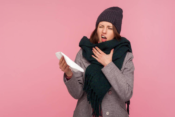 Unhappy sick woman in autumn coat big scarf and warm hat holding napkin and coughing, suffering from seasonal flu. Indoor studio shot isolated on pink background - Фото, зображення