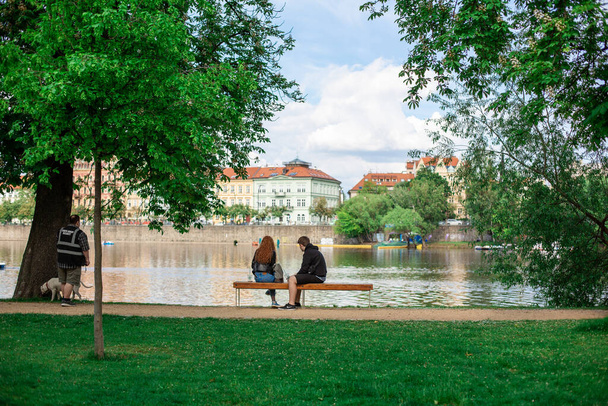 Couple communicate on a bench by the river in a green city park with a city view. Prague / Czechia - 05.21.2019 - Photo, image
