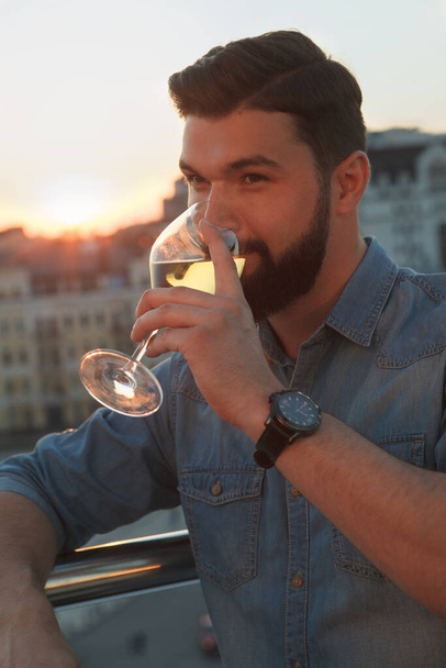 Vertical close up of a bearded man drinking wine on rooftop terrace, watching summer sunset in the city - Foto, imagen