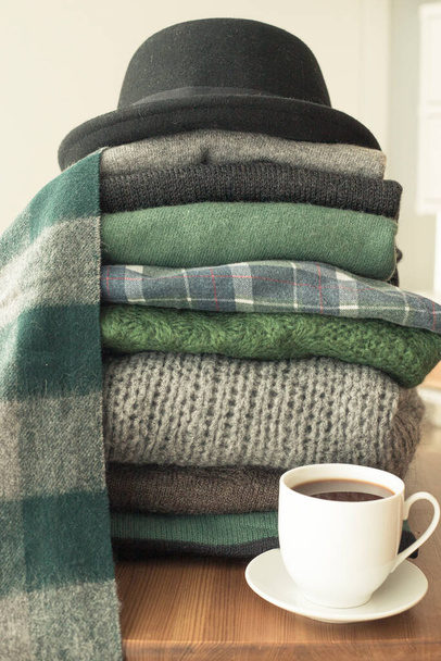 Pile of green and grey sweaters with a black hat on top and a hot drink white cup on the side, background with copy space, cozy, hygge or seasonal wardrobe changing wallpaper. Vertical shot. - Photo, Image