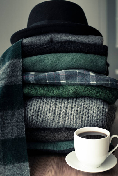Pile of green and grey sweaters with a black hat on top and a hot drink white cup on the side, background with copy space, cozy, hygge or seasonal wardrobe changing wallpaper. Green filter, Vertical shot. - Photo, Image
