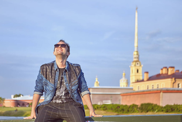 Young man is travelling in Russia. White male tourist in sunglasses is sitting near river in the center of Saint-Petersburg. - Fotoğraf, Görsel