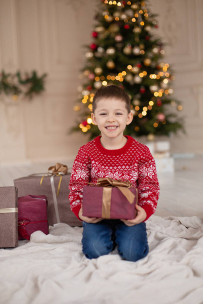 Portrait of happy smiling little boy in red knitted sweater with Christmas present box near Christmas tree. Happy childhood. Waiting for a Christmas tale. - Фото, зображення