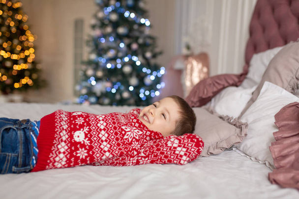 Happy little boy in red knitted sweater, dream on about presents laying in the bed. Christmas tree background. Enjoy winter holidays - 写真・画像