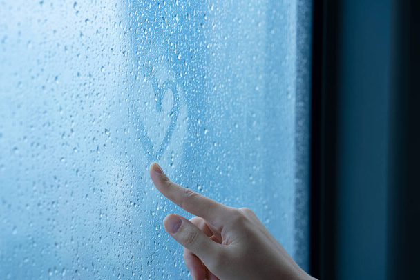 Female hand drawings a heart on a foggy window during the rain. Glass in drops of water. The concept of romance - Foto, Imagen