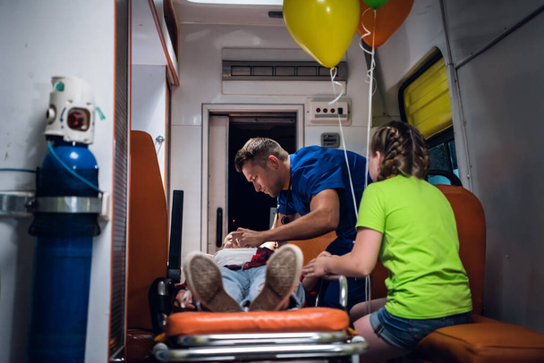 Мать без сознания лежит на носилках в машине скорой помощи, парамедик оказывает ей первую помощь - Фото, изображение