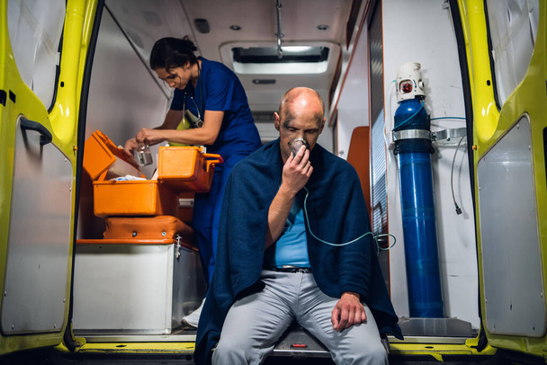 Egy férfi, aki oxigénmaszkban ül egy mentőautóban, egy nővér, aki ellátmányt keres az orvosi felszerelésében. - Fotó, kép
