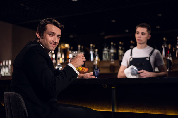 pohledný muž pózuje u baru s koktejlem v ruce na pozadí barmana a lahví - Fotografie, Obrázek