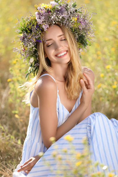Young woman wearing wreath made of beautiful flowers in field on sunny day - Фото, изображение
