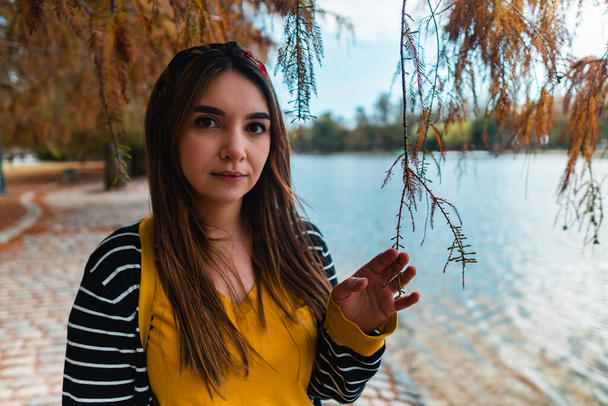 Egy fiatal nő az őszi parkban egy tó mellett, kamerába néz, miközben megérint egy ágat. - Fotó, kép