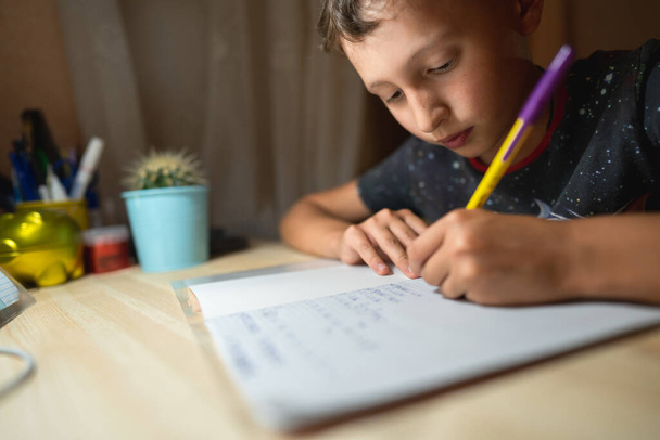 capable schoolboy of 8 years old, writes enthusiastically at the table. The smiling boy is happy to do his homework. Home schooling. - Zdjęcie, obraz