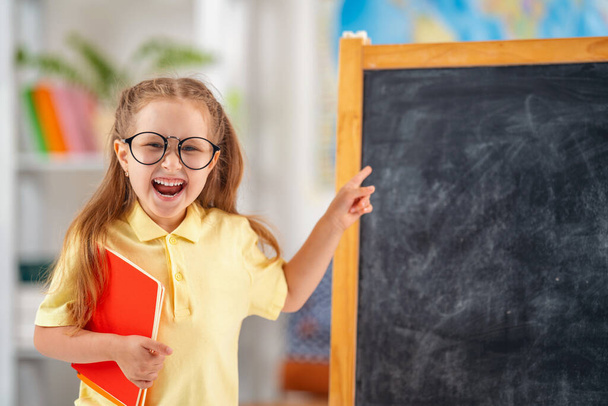 Back to school. funny smiling schoolgirl in glasses with book in her hand, pointing at blackboard. child poses in school classroom against blackboard. Preparation for school. Education. Copy space. - Photo, Image