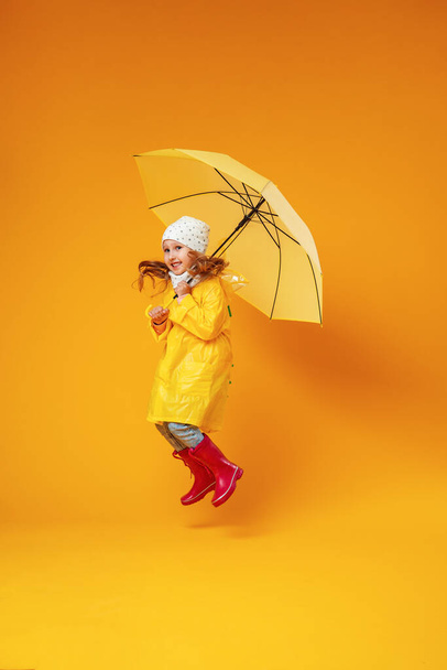 Счастливая эмоционально смешная девочка прыгает и смеется в осенней одежде с желтым зонтиком на цветном желтом фоне - Фото, изображение