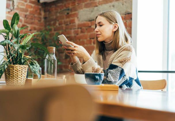 Молодая блондинка в повседневной одежде смотрит на смартфон во время обеда в кафе - Фото, изображение