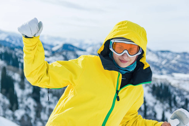 Mooie youg vrouw toerist in gele jas, skibril masker en witte wanten op een achtergrond van bergen kijken naar de camera. Wintervakantie en tijdverdrijf concept. - Foto, afbeelding