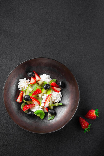 Bord met fruitsalade, kiwi, aardbeien, bosbessen, appel, slagroom - Foto, afbeelding