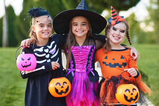 Optimistic girls in creepy costumes smiling for camera and embracing each other while trick or treating in par - 写真・画像