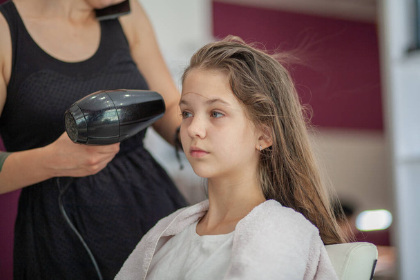 Beautiful young European girl sits in a beauty salon, professional hairdresser stylist works with long hair of a girl. - Photo, Image