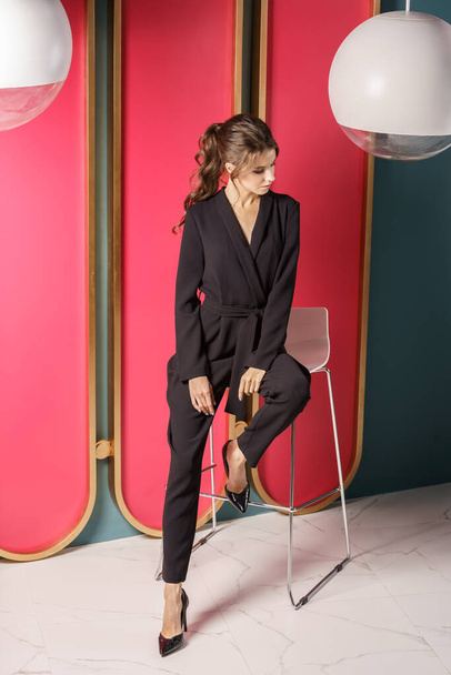 Beautiful businesswoman standing close to red wall at luxury apartments interior. Portrait of beauty brunette in black suit and high heels  - Φωτογραφία, εικόνα