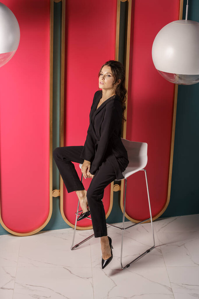 Beautiful businesswoman posing close to red wall at luxury apartments interior. Portrait of beauty brunette in black suit and high heels  - 写真・画像