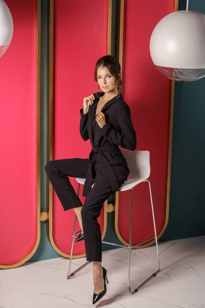 Beautiful businesswoman posing close to red wall at luxury apartments interior. Portrait of beauty brunette in black suit and high heels  - Fotoğraf, Görsel