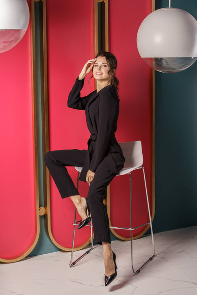 Beautiful businesswoman posing close to red wall at luxury apartments interior. Portrait of beauty brunette in black suit and high heels  - Foto, Bild