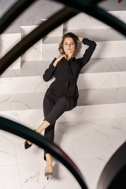 Beautiful businesswoman posing on white stairs at luxury apartments interior. Portrait of beauty brunette in black suit and high heels  - 写真・画像