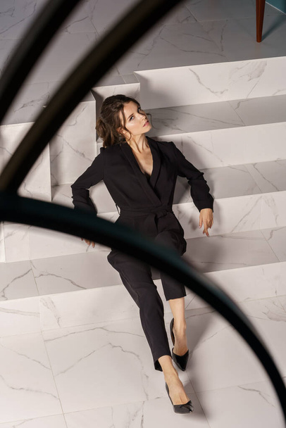 Beautiful businesswoman posing on white stairs at luxury apartments interior. Portrait of beauty brunette in black suit and high heels  - Zdjęcie, obraz