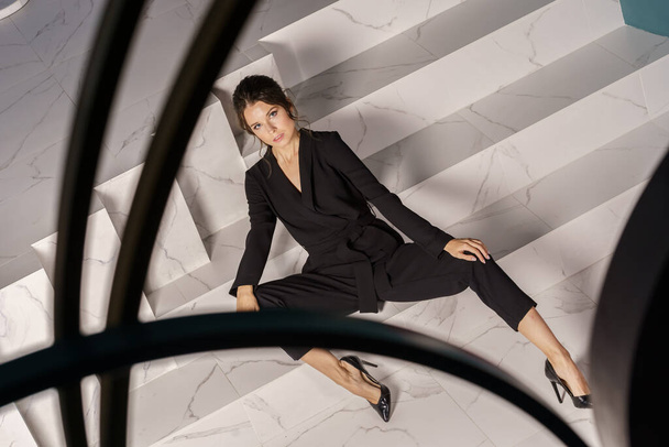 Krásná podnikatelka pózující na bílých schodech v luxusních apartmánech interiéru. Portrét krásy brunetky v černém obleku a vysoké podpatky  - Fotografie, Obrázek
