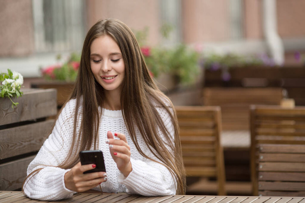 Jovem mulher bonita em um café à espera de uma ordem. Smartphone em suas mãos e um sorriso - Foto, Imagem