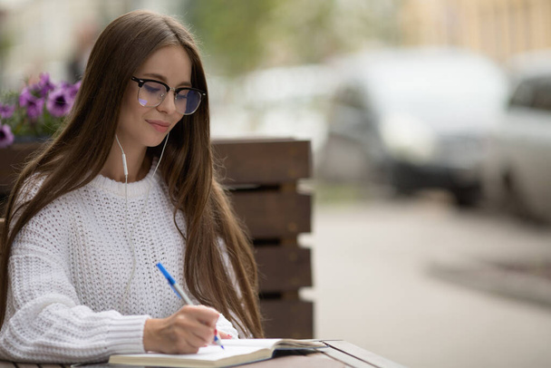 donna urbana seduta in un caffè con occhiali e cuffie. Scrivere in un quaderno - Foto, immagini