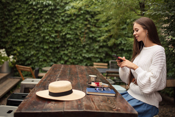 Bir kafede gözlüklü ve kulaklıklı bir kadın oturuyor. Elinde telefonla müzik dinlersin. İş yemeği, kahve ve peynirli kek. - Fotoğraf, Görsel