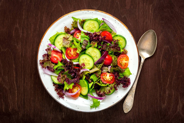 Здоровый овощной салат из помидоров черри, огуречных ломтиков, зеленых и фиолетовых листьев салата, лука и оливкового масла в тарелке на деревянном столе. - Фото, изображение