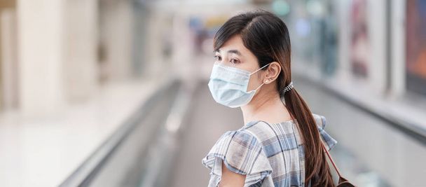Young adult female wearing face mask in airport terminal, protection Coronavirus disease infection, Asian woman traveler walking on escalator in aerodrome. New Normal and travel under COVID-19 concept - Photo, image