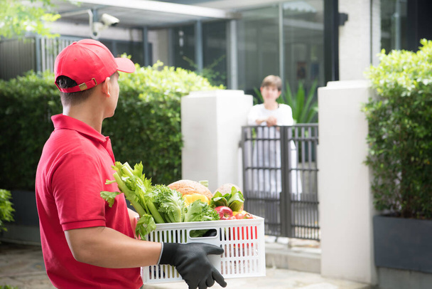 aziatische levering man in rood uniform leveren boodschappen doos van voedsel, fruit, groente en drinken naar vrouw ontvanger  - Foto, afbeelding
