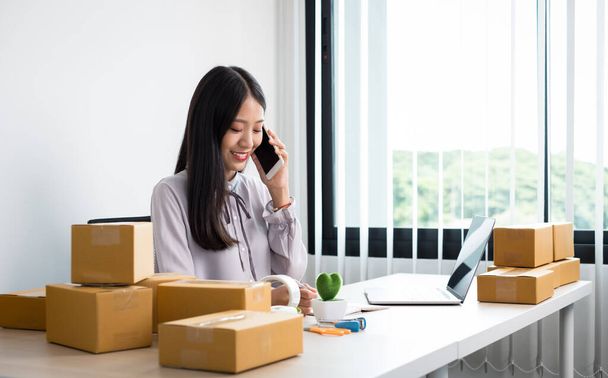 Young Asian woman business owner with many parcel boxes on the table happy online sales job, use your smartphone, get an order from customers, take notes, and make arrangements for delivery by post. - Photo, Image