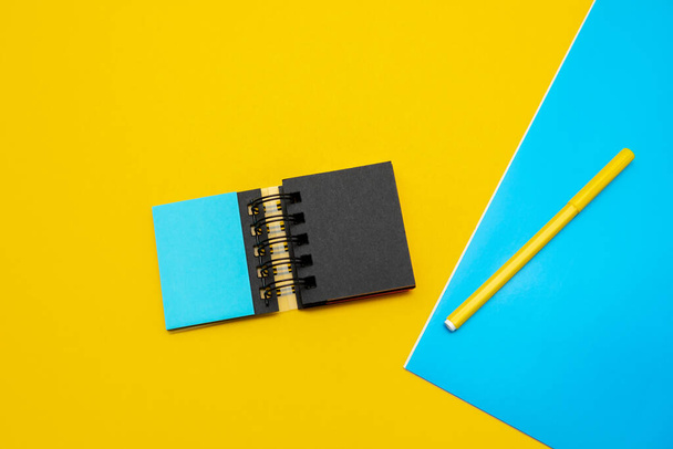 黄色の背景にオフィス用品。2色のノートパッド、オレンジのペン、そして青い段ボールのピース。文房具のコンセプト. - 写真・画像