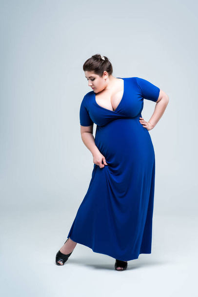 Plus size fashion model in blue dress with deep neckline, fat woman on gray studio background, body positive concept - 写真・画像