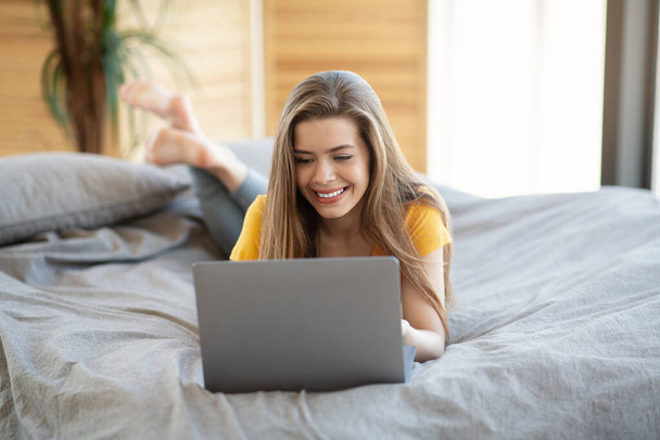 Hübsches Millennial-Mädchen mit Laptop, das online arbeitet oder lernt, während es zu Hause auf seinem Bett liegt - Foto, Bild