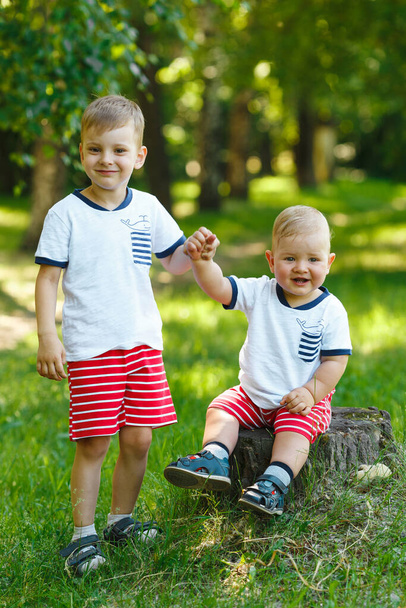 Дети в зеленом летнем лесу. Два младших брата на деревянном пне в весеннем парке. Семейная прогулка на открытом воздухе. Дружеские отношения в семье. Та же одежда. - Фото, изображение