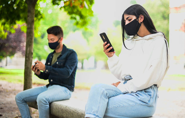 A fiatalok távolságot tartanak a koronavírus miatt, miközben telefonjukat a szabadban használják egy parkban. - Fotó, kép
