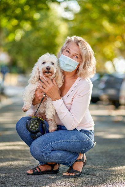 Mujer madura paseando con un perro al aire libre una máscara antivirus. - Foto, imagen