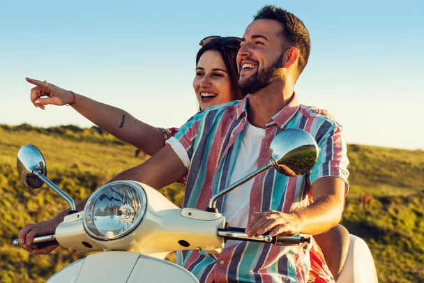 Pareja de jóvenes amigos con ropa fresca, divirtiéndose y sonriendo en una motocicleta en la costa durante la luz del atardecer - Foto, imagen