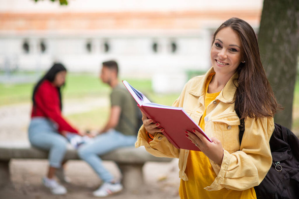 Woman student holding a book in front a a group of friends in park - Photo, image