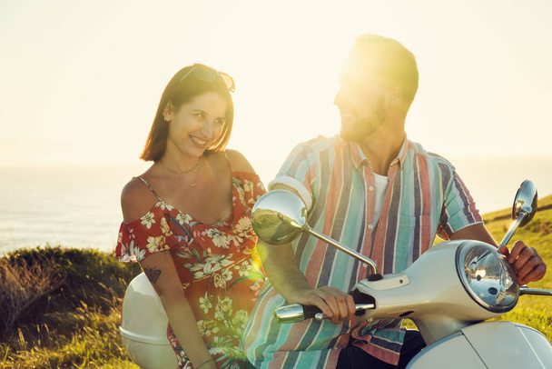 Δύο νεαροί φίλοι με φρέσκα ρούχα, διασκεδάζουν και χαμογελούν σε μια μοτοσικλέτα στην ακτή κατά τη διάρκεια του ηλιοβασιλέματος - Φωτογραφία, εικόνα