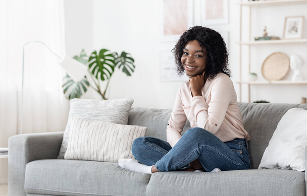 Shy Black Girl Sitting On Sofa At Home, Posing To Camera - 写真・画像
