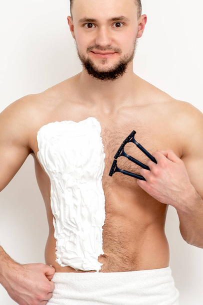 Young caucasian man with beard holds razor shaves his chest with white shaving foam on white background. Man shaving his torso - Photo, Image