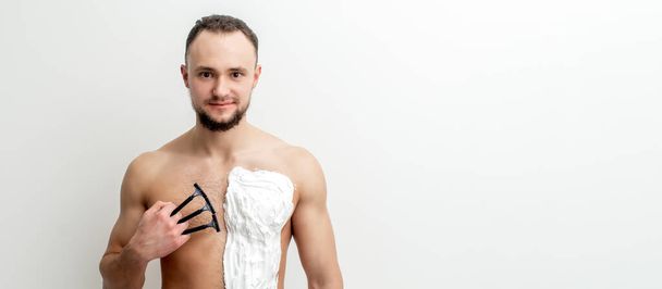 Junger kaukasischer Mann mit Bart rasiert sich mit weißem Rasierschaum auf weißem Hintergrund die Brust. Mann rasiert sich Oberkörper - Foto, Bild