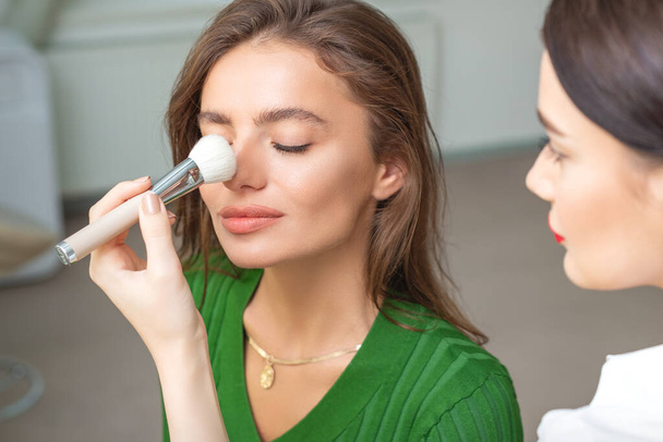 Sminkes alkalmazó száraz kozmetikai tónus alapja orra fiatal kaukázusi nő ecsettel eszköz szépségszalonban - Fotó, kép
